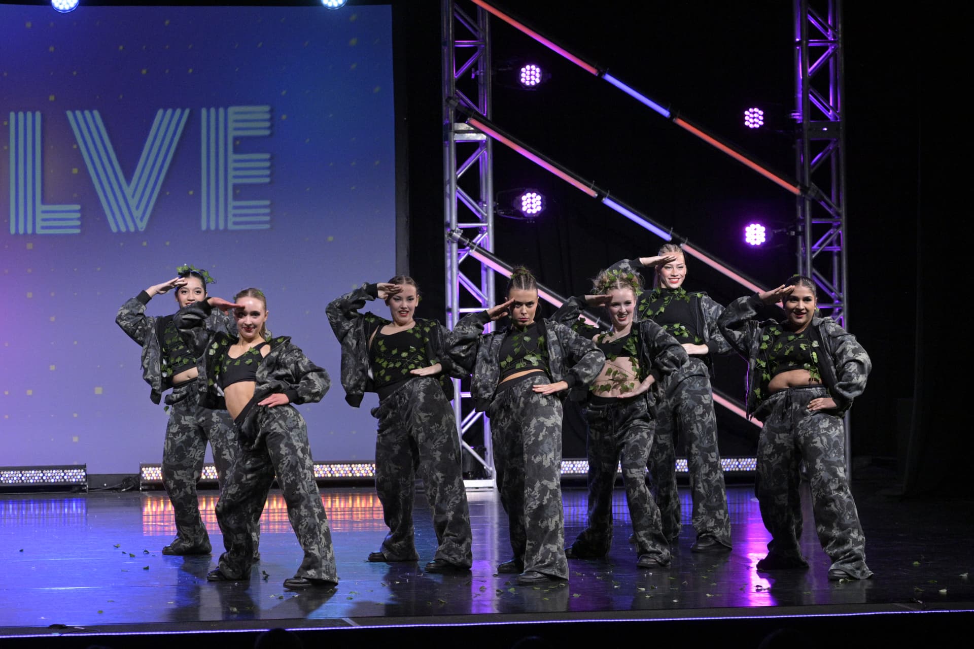 Seven Danscott hip hop dancers in camouflage outfits, saluting in a staggered line on a stage lit in purple