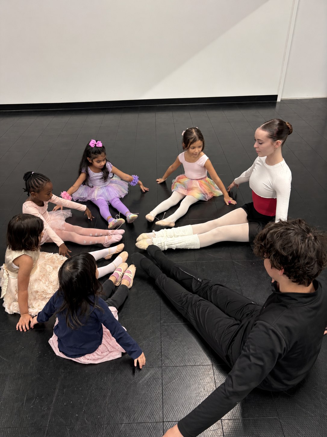 Young dancers sitting in a circle on the studio floor with two teachers