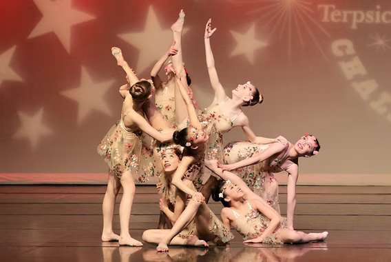 Danscott lyrical dancers in nude costumes with floral skirts, arranged in a sculptural group pose on a stage lit with a red gradient and white stars